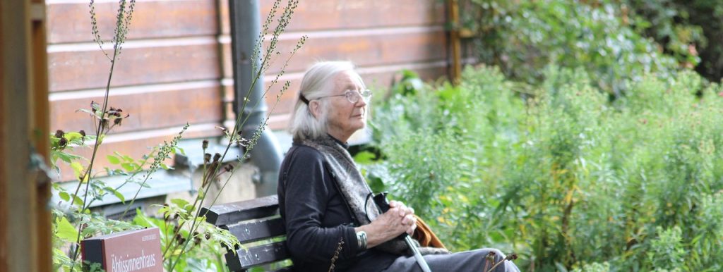 Older woman sitting on a bench beside a brick building and garden