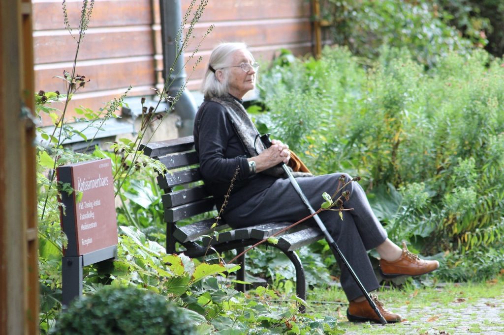 Older woman sitting on a bench beside a brick building and garden